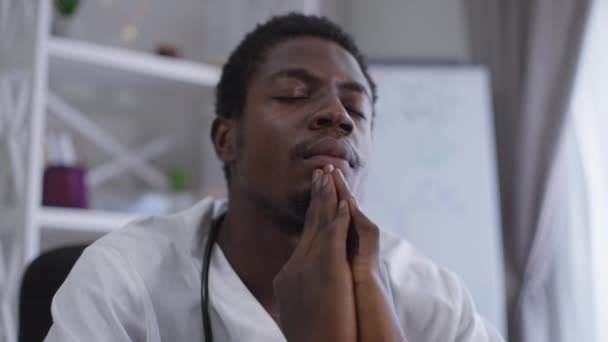 Junger afroamerikanischer Arzt betet mit geschlossenen Augen im Krankenhaus. Porträt des Hoffnungsträgers bei Ausbruch der Coronavirus-Pandemie am Arbeitsplatz. Gesundheitsversorgung und Glaubenskonzept. — Stockvideo