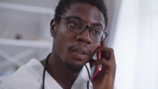 Close-up positivo médico confiante em óculos falando no telefone sorrindo. Retrato de jovem afro-americano consultando paciente em pé dentro do hospital. — Vídeo de Stock