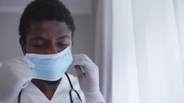 Médico varón en guantes quirúrgicos que se pone la mascarilla del coronavirus de pie en el hospital en interiores. Primer plano retrato serio hombre afroamericano preparándose por la mañana en la pandemia de Covid-19. — Vídeos de Stock