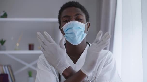 Vista frontal retrato serio médico afroamericano en Covid mascarilla gesto no cruzar las manos mirando a la cámara. Experto profesional joven posando en el interior del hospital sobre la pandemia de coronavirus. — Vídeo de stock