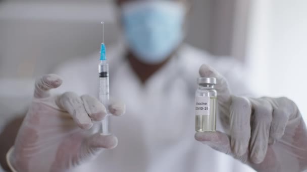 Close-up syringe and ampoule with coronavirus vax in male African American hands in gloves. Blurred man doctor showing Covid-19 treatment posing indoors in hospital. Illness prevention concept. — Stock Video