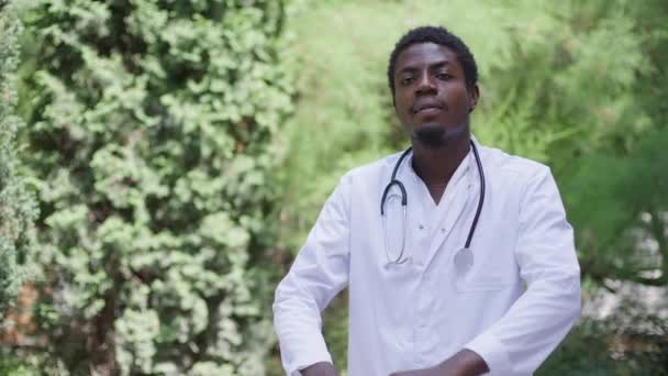 Självsäker glad snygg afroamerikansk man i läkaruniform som korsar händerna och tittar på kameran stående i parken utomhus. Porträtt av expert professionell läkare poserar i slow motion. — Stockvideo