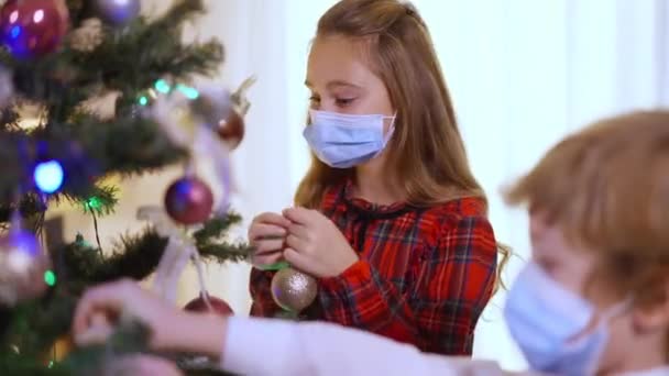 Veselá běloška v koronavirové masce zdobí vánoční stromek s bratrem v obývacím pokoji doma. Portrét pozitivní sestry a chlapce připravující dům na novoroční oslavu na Covid-19 — Stock video