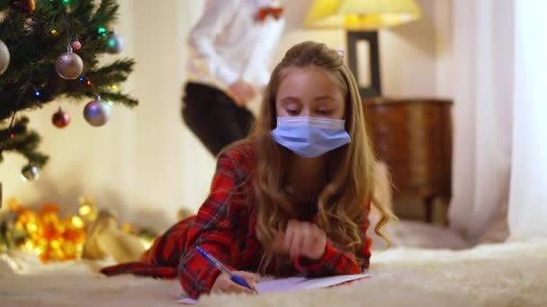 Soustředěná dívka v Covid-19 obličejové masky psaní dopis Santa Clause jako chlapec vstupující do obývacího pokoje mluvit. Portrét roztomilých bělochů v obývacím pokoji doma na Štědrý večer.