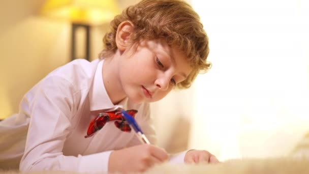 Fokuserad charmig pojke skriva brev till jultomten tittar på kameran leende. Porträtt av glada kaukasiska barn liggande på mjuk matta med penna tänkande poserar inomhus på nyårsafton. — Stockvideo