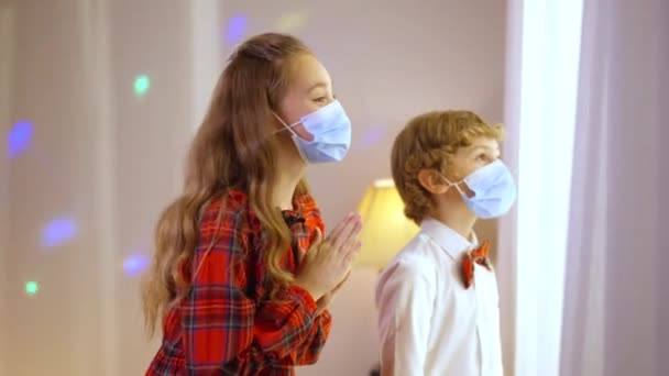 Side view excited Caucasian boy and girl in coronavirus face masks gesturing thumbs up smiling waiting for Santa looking out the window. Happy excited brother and sister at home indoors on New Year. — Stock Video