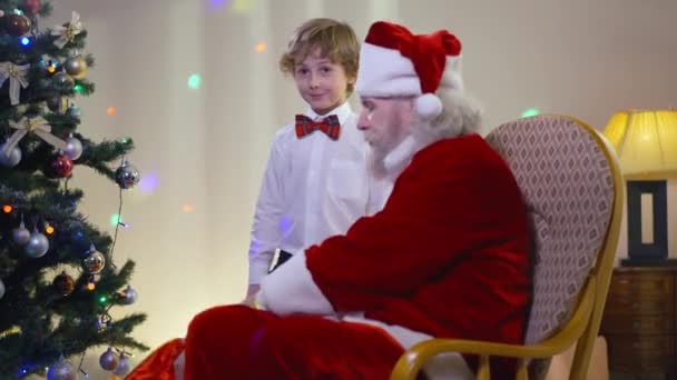 Porträt eines aufgeregten kleinen Jungen, der an Neujahr Geschenke erhält und den Weihnachtsmann im Schaukelstuhl umarmt. Glückliche süße kaukasische Kind überrascht mit Geschenk umarmt Weihnachtsmann zu Hause lächelnd. — Stockvideo