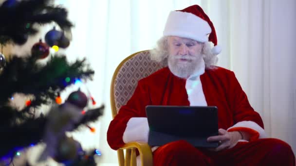 Noel Baba 'nın sallanan sandalyede oturup tablet üzerinde internette sörf yaptığı pozitifti. Oturma odasındaki Noel ağacında dişlek gülüşü olan mutlu, yaşlı, beyaz sakallı adam.. — Stok video