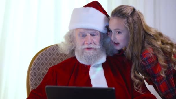 Porträt des alten Weihnachtsmannes mit Tablet als fröhliches kleines Mädchen, das am Ohr flüstert. Lächelnder alter bärtiger Kaukasier im roten Kostüm gestikuliert mit dem Daumen nach oben als Kind, das sich drinnen wünscht. Neujahrsfeier. — Stockvideo