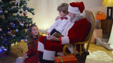 Noel Baba 'yla konuşan büyüleyici kız ve oturma odasındaki yeni yıl ağacında sallanan sandalyede oturan çocuk. Kafkasyalı mutlu çocuklar Noel Baba 'nın evde kutlanmasının tadını çıkarıyor..