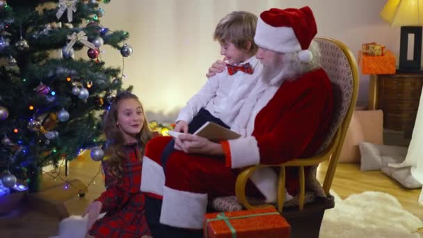 Podekscytowana urocza dziewczyna rozmawiająca z Ojcem Bożym i chłopcem siedzącym na bujanym krześle w Nowym Roku w salonie. Szczęśliwe dzieci kaukaskie korzystających święto wakacje z Santa Clause w pomieszczeniach. — Wideo stockowe