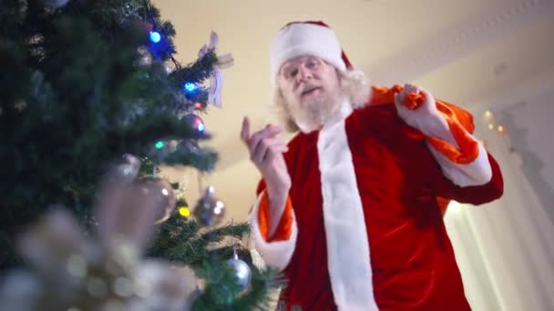 Neşeli Noel Baba, hediye çantasıyla kameraya eğiliyor. Dudaklarına parmak işareti yapıyor, gülümsüyor. Noel arifesinde oturma odasındaki Noel ağacında kostümlü neşeli, sakallı, beyaz bir adamın portresi.. — Stok video