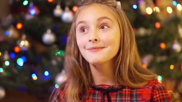 Kopfballporträt eines aufgeregten charmanten kaukasischen Mädchens, das sich an Heiligabend ein Wunder wünscht, während im Hintergrund der geschmückte Neujahrsbaum funkelt. Close-up glückliches Kind drinnen im Urlaub. — Stockvideo