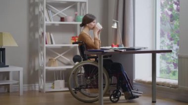 Yan görünüm güzel beyaz kadın tekerlekli sandalyeli engelli sabah kahvesi içen camdan dışarı bakan. Geniş açılı, kendinden emin genç serbest çalışan ev ofisinde sıcak içkinin tadını çıkarıyor..