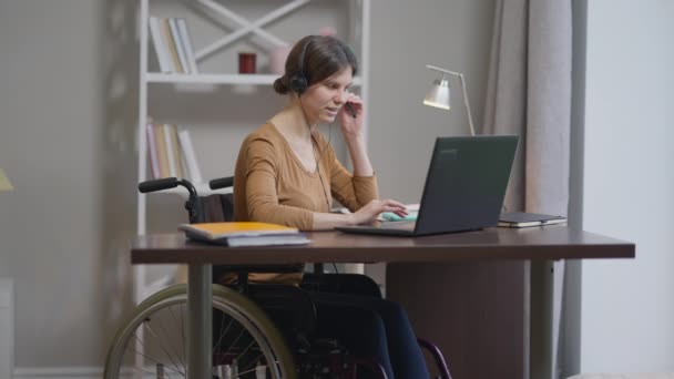 Smiling confident professional disabled freelancer in headphones talking in slow motion texting on laptop keyboard conferencing online. Portrait of young Caucasian woman messaging on remote working. — Stock Video