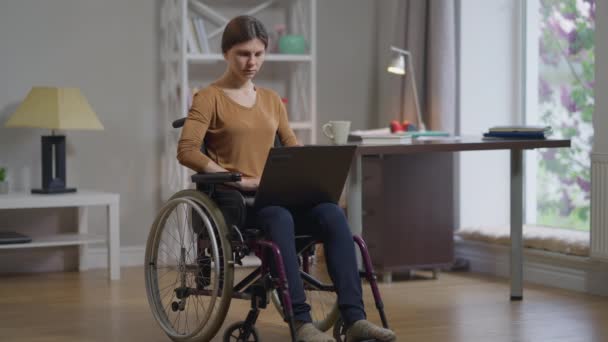 全景拍摄严重残疾的年轻女子坐在轮椅上，膝上型电脑在网上输入键盘讯息。浓缩白人美女自由职业者慢镜头上网的肖像. — 图库视频影像