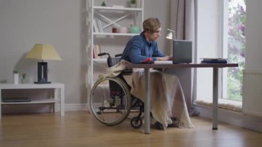 Geniş açılı portre, akıllı, kendine güvenen, tekerlekli sandalyedeki özürlü, dizüstü bilgisayarlı ve evraklı adam. Yavaş çekimde çalışan odaklanmış genç serbest çalışan..
