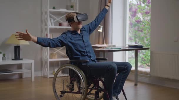 Smiling happy disabled man in VR headset enjoying augmented reality sitting in wheelchair at home. Wide shot joyful Caucasian handicapped guy travelling online using modern technologies. — Stock Video