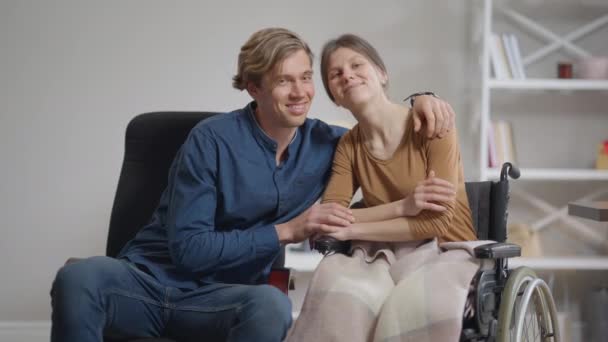 Front view portrait happy hugging Caucasian woman in wheelchair and man watching TV talking in slow motion. Cheerful loving couple indoors at home on weekend enjoying leisure. Disability and love. — Stock Video