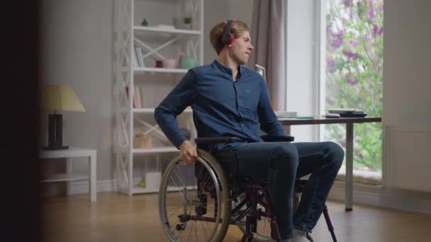 Happy Caucasian disabled man dancing in wheelchair listening to music in headphones. Wide shot positive cheerful paralyzed guy enjoying hobby at home indoors in slow motion. Lifestyle individuality. — Stock Video