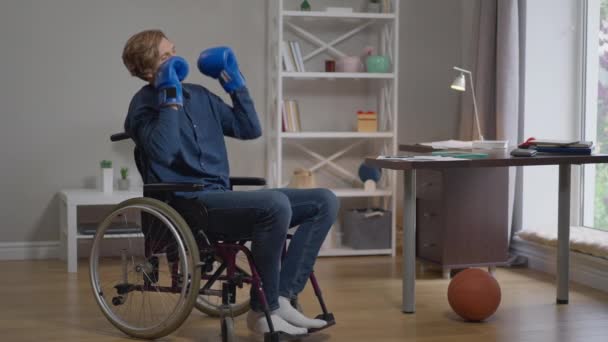 Jonge sporter in een rolstoel die in slow motion bokshandschoenen aantrekt die tegen de schaduw vechten. Brede shot portret gemotiveerde blanke gehandicapte man training binnen thuis. Paralympisch sportconcept. — Stockvideo