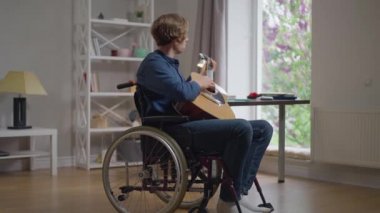 Evde tekerlekli sandalyede oturan, gitar çalan yetenekli, engelli beyaz adam portresi. Rahat, yetenekli gitarist yavaş çekimde hobisinin tadını çıkarıyor. Yetenek ve yaşam tarzı konsepti.