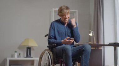 Mutlu engelli adam yavaş çekimde akıllı telefondan video oyunu oynuyor. Tekerlekli sandalyedeki gülümseyen beyaz adamın portresi evde keyfine bakıyor..