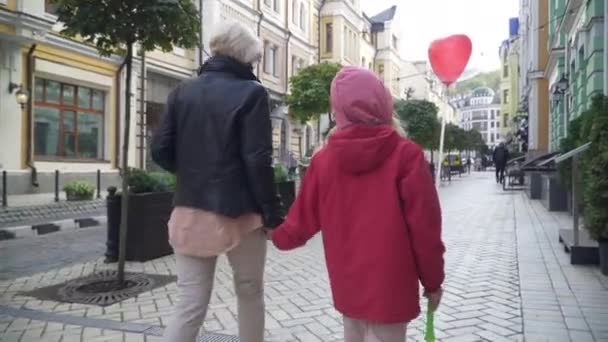 Vista posterior Abuela caucásica en coronavirus mascarilla caminar con la nieta en la calle de la ciudad hablando. Feliz abuela y chica paseando al aire libre disfrutando de fin de semana en la pandemia de Covid-19. — Vídeo de stock