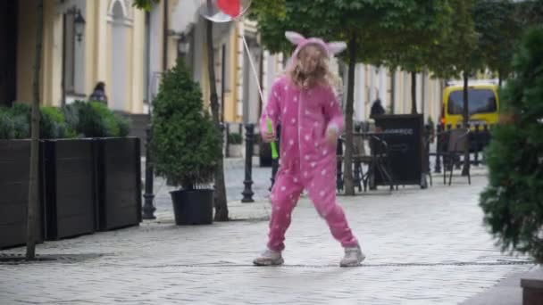 Ευρεία shot πορτρέτο χαρούμενος ενθουσιασμένος Καυκάσιος κορίτσι σε ροζ κοστούμι με μπαλόνι άλμα χαμογελώντας κοιτάζοντας κάμερα. Χαρούμενο παιδί που διασκεδάζει στο δρόμο της πόλης το Σαββατοκύριακο απολαμβάνοντας ελεύθερο χρόνο. — Αρχείο Βίντεο