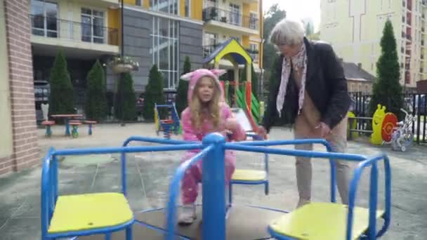 Sonriente hermosa abuela balanceándose carrusel con linda nieta sentada en la plataforma giratoria. Amplio retrato retrato feliz mujer caucásica y niño disfrutando de ocio en el patio de recreo. — Vídeos de Stock