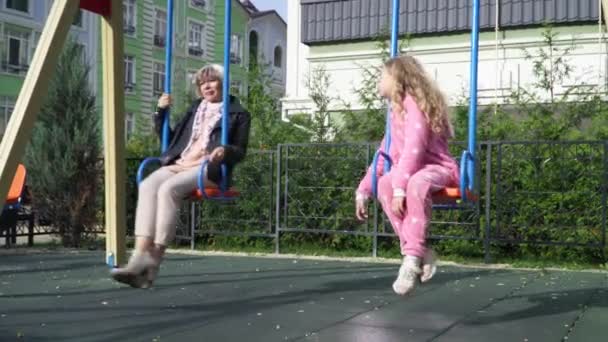 Wide shot portrait little girl and senior woman swinging on swings on sunny day in city talking. Relaxed Caucasian granddaughter and grandmother enjoying leisure outdoors on playground chatting. — Stock Video