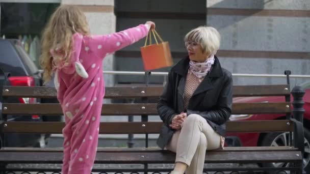 Charmante schattige kleindochter verrassende oma met cadeau op zonnige stad straat. Portret van een vrolijke glimlachende blanke oudere vrouw die een mooi meisje knuffelt. Vrije tijd en levensstijl. — Stockvideo