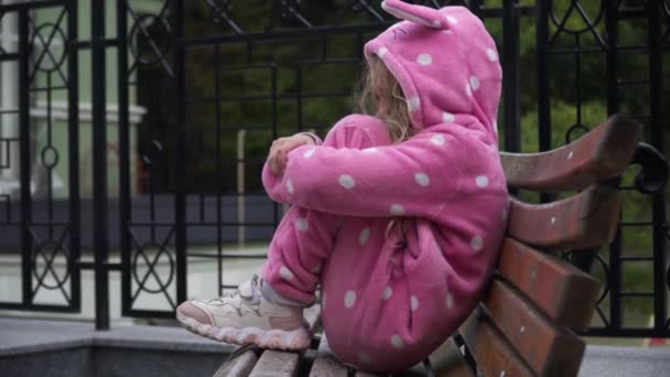 Vista lateral triste chica solitaria en traje rosa sentado en el banco al aire libre pensando. Retrato de un niño caucásico solo en la calle de la ciudad. Tristeza y concepto infantil. — Vídeo de stock