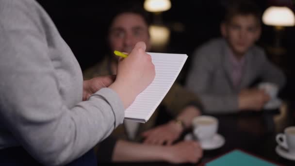 Close-up mão garçonete irreconhecível escrever com caneta em câmera lenta recebendo ordem no restaurante de clientes desfocados sentados à mesa. Profissional caucasiano jovem servindo pessoas no café. — Vídeo de Stock