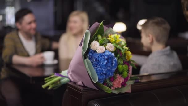 Närbild vacker bukett blommor i restaurang med suddig glad kaukasisk familj talar sitter vid bordet i bakgrunden. Avslappnad man kvinna och pojke njuter av middag på café på semester. — Stockvideo