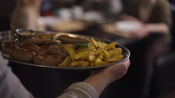 Cámara en vivo sigue bandeja con patatas a la parrilla y salchichas en manos del camarero caminando a la mesa con los clientes. Empleado caucásico irreconocible sirviendo comida en el restaurante en cámara lenta. — Vídeo de stock