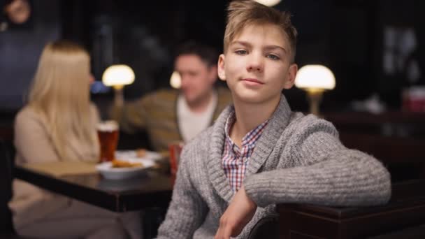 Feliz adolescente orgulloso mirando a la cámara sonriendo con los padres borrosos hablando de fondo. Retrato de adolescente caucásico positivo posando en el interior cenando con el padre y la madre. Movimiento lento. — Vídeos de Stock