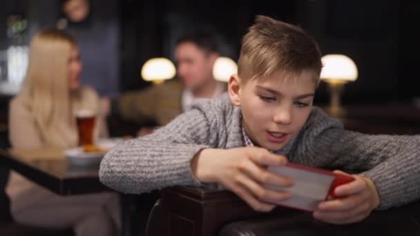 Absorbido adolescente haciendo gestos de victoria en los juegos de cámara lenta en línea en el teléfono inteligente. Pareja borrosa de padres sentados en el restaurante hablando de fondo. Caucásico adolescente jugando videojuegos. — Vídeo de stock