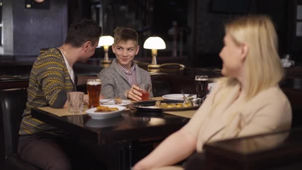 Rack fokussiert freudige Vater und Teenager-Sohn, die Spaß dabei haben, lächelnd im Restaurant zu einer verlegenen Frau vor der Kamera mit unzufriedenem Gesichtsausdruck zu sprechen. Individualitätskonzept. — Stockvideo