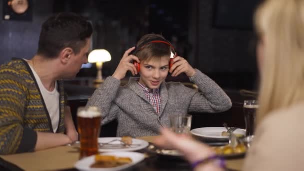 Se comporter mal adolescent fils mettre des écouteurs comme des parents gronder garçon assis dans le restaurant. Adolescent caucasien malheureux avec père et mère dînant dans un café à l'intérieur. Mouvement lent. — Video