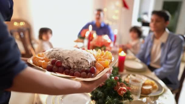 Vaschetta da primo piano con deliziosa pasticceria natalizia e frutta in mano femminile con sfocata famiglia mediorientale che applaude seduta a tavola sullo sfondo. Felice famiglia amorevole che celebra il nuovo anno a casa. — Video Stock