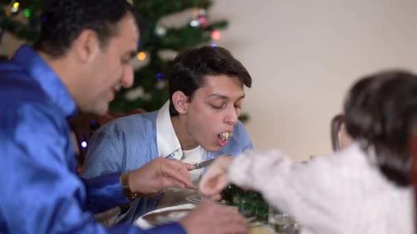 Sonriente adolescente chico de Oriente Medio disfrutando del sabor de la pastelería dulce en Navidad sentado en la mesa con la familia. Feliz hijo celebrando el Año Nuevo con el padre y el hermano pequeño en casa. Concepto familiar. — Vídeos de Stock