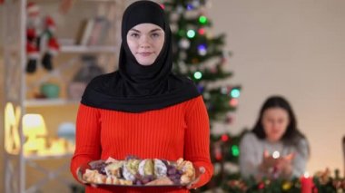 Kafiyeli genç ve mutlu bir kadın evdeki oturma odasında lezzetli bir Noel tatlısıyla poz veriyor. Arka planda bulanık bir arkadaş oturuyor. Gururlu ev kadınının portresi Yeni Yıl ekmeğiyle gülümseyen kameraya bakıyor.