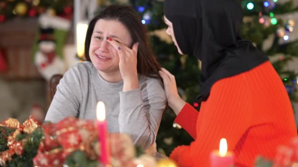 Retrato de llorando frustrada mujer joven caucásica sentada en la mesa de Navidad con un amigo musulmán en hiyab calmándose animando a hablar. Deprimida dama apoyada en Año Nuevo en casa. — Vídeos de Stock