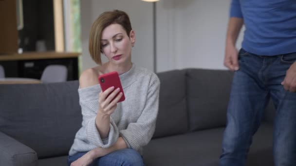 Schöne Frau, die per Smartphone SMS schreibt und dabei den Mann ignoriert, der sich mit schockiertem Gesichtsausdruck auf die Couch setzt. Porträt einer betrügerischen kaukasischen Ehefrau, die mit Liebhaber und Ehemann kommuniziert. — Stockvideo