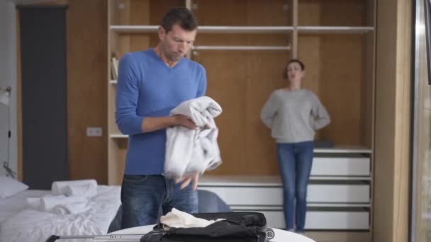 Sorgsen make packar resväska i sovrum med suddig fru pratar i bakgrunden. Porträtt av en vuxen vit man som lämnar en stressad kvinna. Begreppet relationssvårigheter och skilsmässa. — Stockvideo