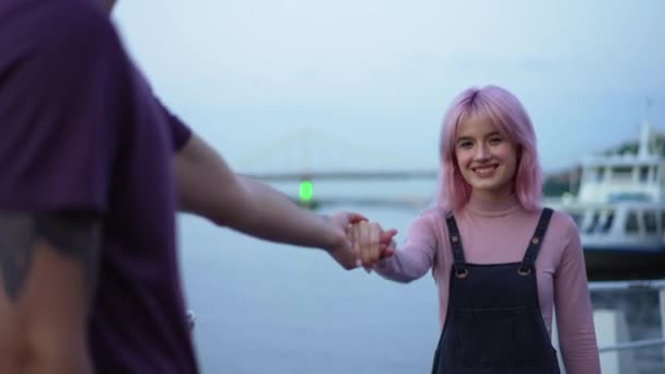 Young confident smiling woman with pink hair and nose piercing walking holding male hand looking at camera. Portrait of cheerful happy Caucasian girlfriend dating with boyfriend on urban embankment. — Stock Video