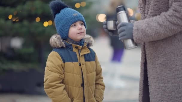 Retrato de menino bonito do Oriente Médio em pé na árvore de Natal na cidade com mulher irreconhecível derramando chá quente de garrafa térmica. Filho feliz desfrutando feriados de Ano Novo com a mãe ao ar livre. — Vídeo de Stock