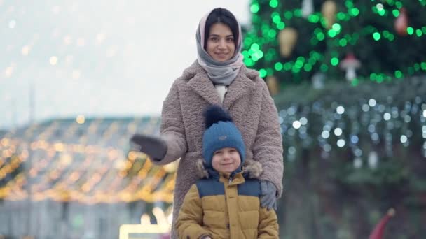 Magabiztos közel-keleti gyönyörű nő és aranyos fiú integetett a kamerába álló szilveszterkor fa a város főterén. Boldog anya és fia portréja a szabadban karácsony este. — Stock videók