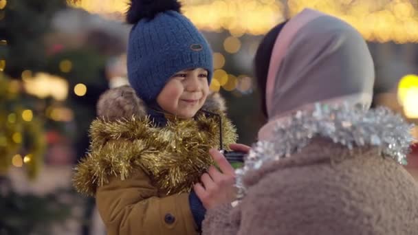Sød spændt søn taler med mor på byens torv dekoreret til jul. Portræt af positiv glad mellemøstlig dreng nyder nytårsferie fest med kvinde udendørs. – Stock-video